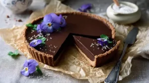 Tarte au chocolat sans cuisson avec base biscuitée dorée et ganache lisse, décorée d’une fleur violette.
